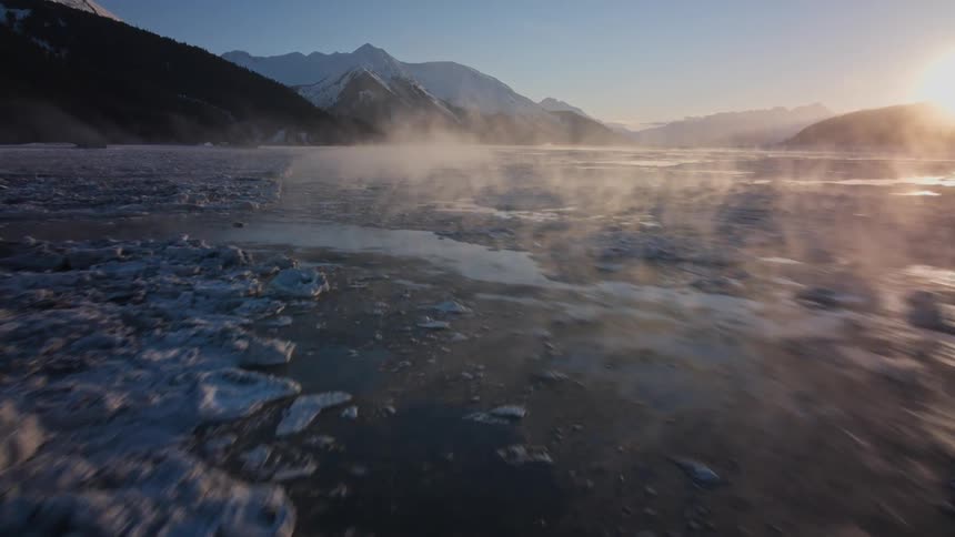 Frost rises from a cold lake below a mountain range