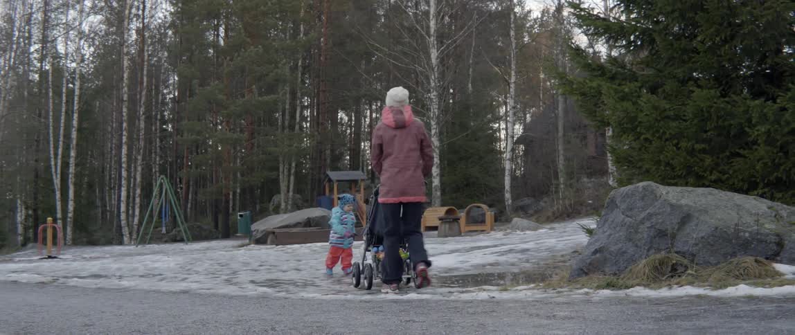 A woman pushes a stroller in a snow covered park while her toddler plays