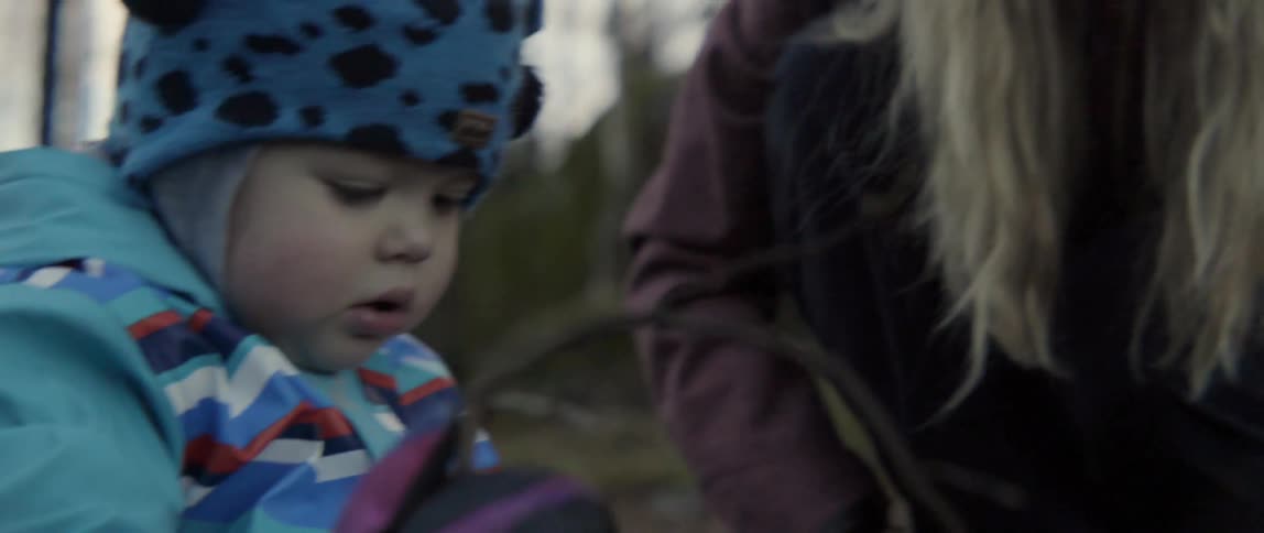 A baby holds a twig while sitting in a forested area on a chilly day