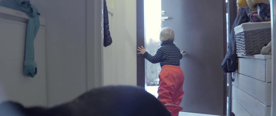 A toddler wearing red snow pants pushes open a door to go play outside.