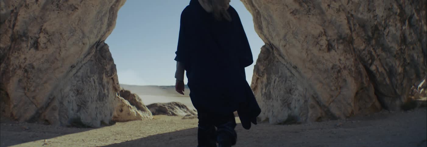 An elderly woman walks towards a rock formation in a desert during the day