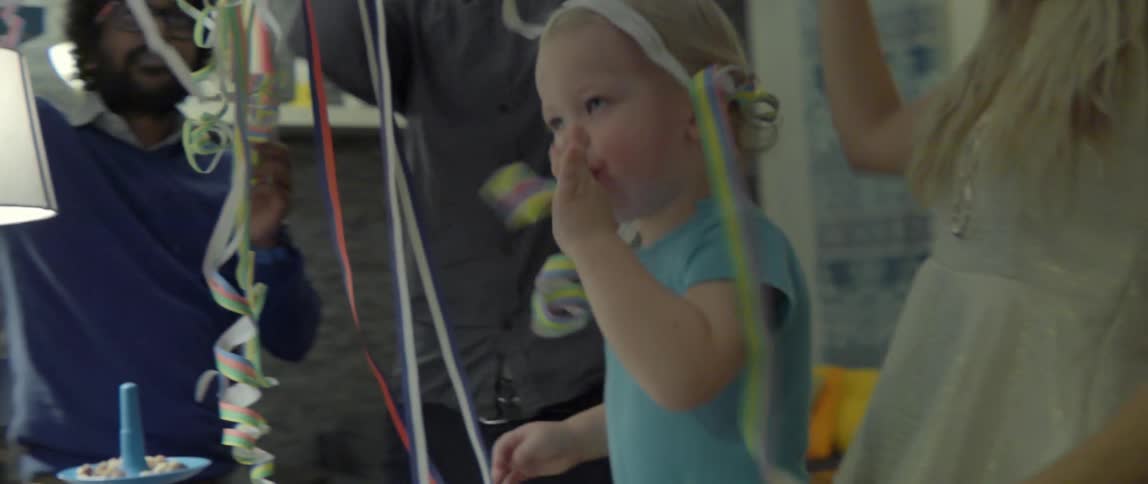 A young toddler blowing a colorful party horn while surrounded by festive paper streamers.