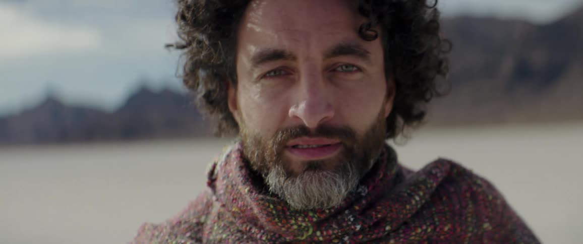 A man looks into the camera as he comes to a stop before it in a desert during the day