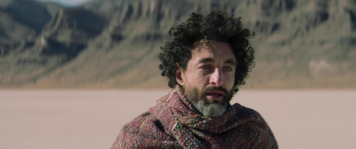 A man walks backwards in a desert and then wipes a tear away and turns around and walks away