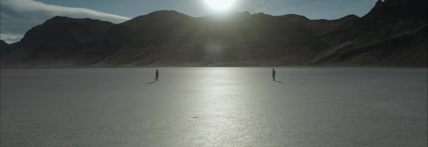 Two people silhouetted by the sun approach each other across desert sand with hills in the background