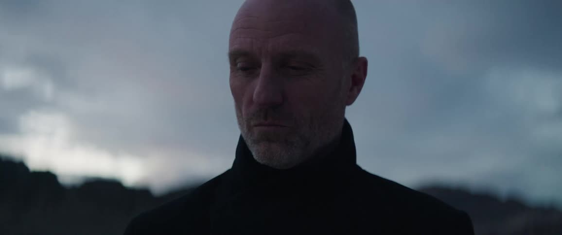 A bald man standing outside on a cloudy, cold evening