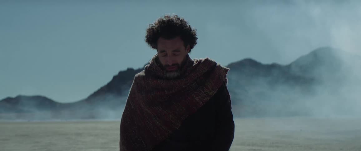 A man in a desert kneels in the sand and prays