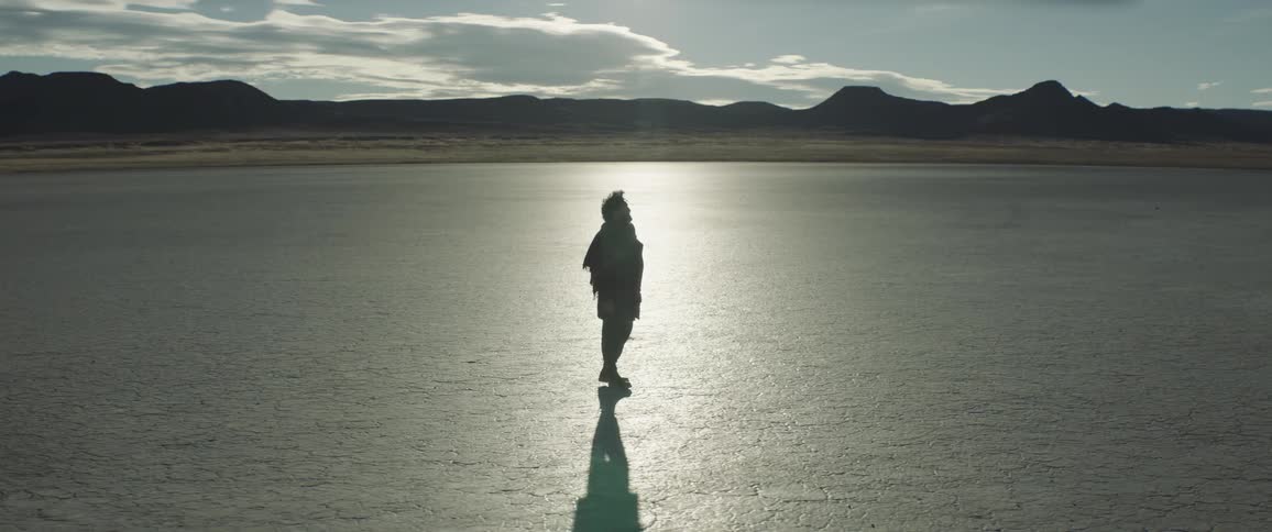 A man walking on cracked earth flats stops to yell and search then continues on