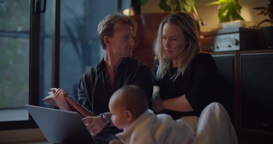 A man and woman sit on the floor at home and look at a book and laptop as their baby daughter reaches over to touch the screen