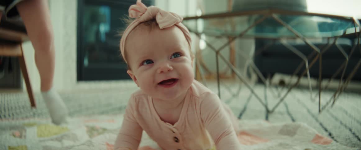 A smiling baby girl with her sibling playing in the background