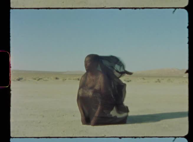 A woman covered in a body length veil kneels in an arid flatland on a windy day