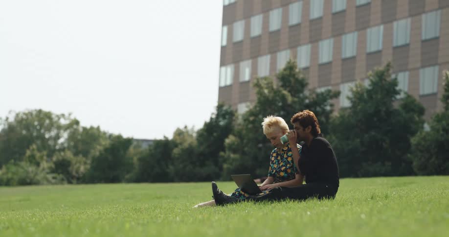 A teenage uses a laptop as she sits next to a boy on a hill who is drinking a coffee