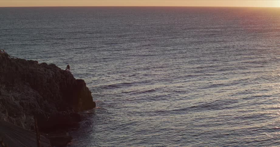 Fisherman stands on cliff overlooking ocean at sunset