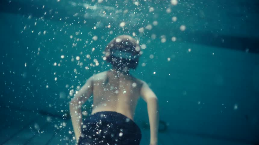 A boy swimming underwater in a pool heads back to the sunny surface