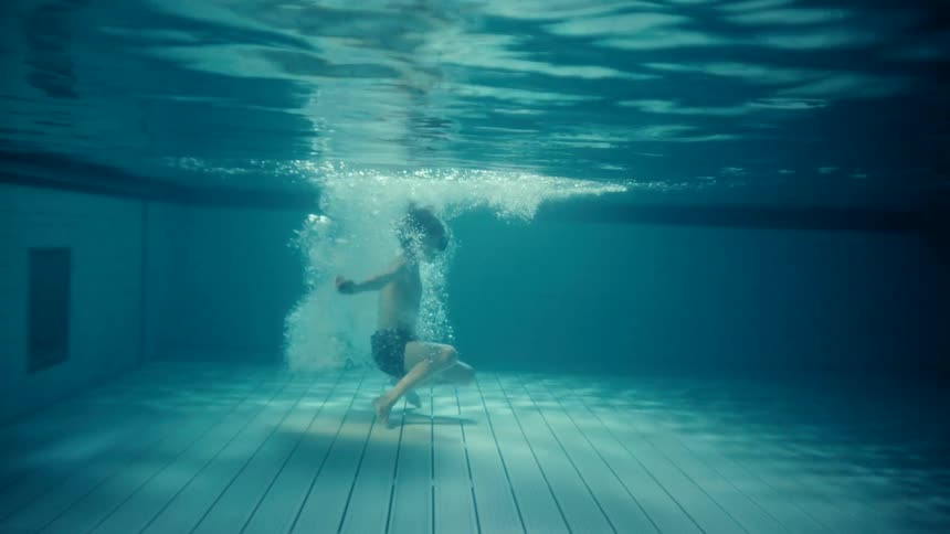 Sunlight shines on the water in a swimming pool as a little boy jumps in and lands on the bottom before he swims away