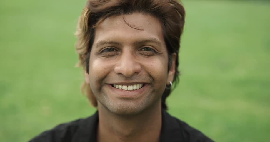 A man is smiling and sitting outdoors in a grass field