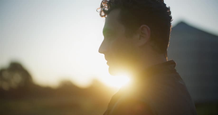 Man slightly silhouetted against the setting sun stands near a silo and manipulates a drone controller