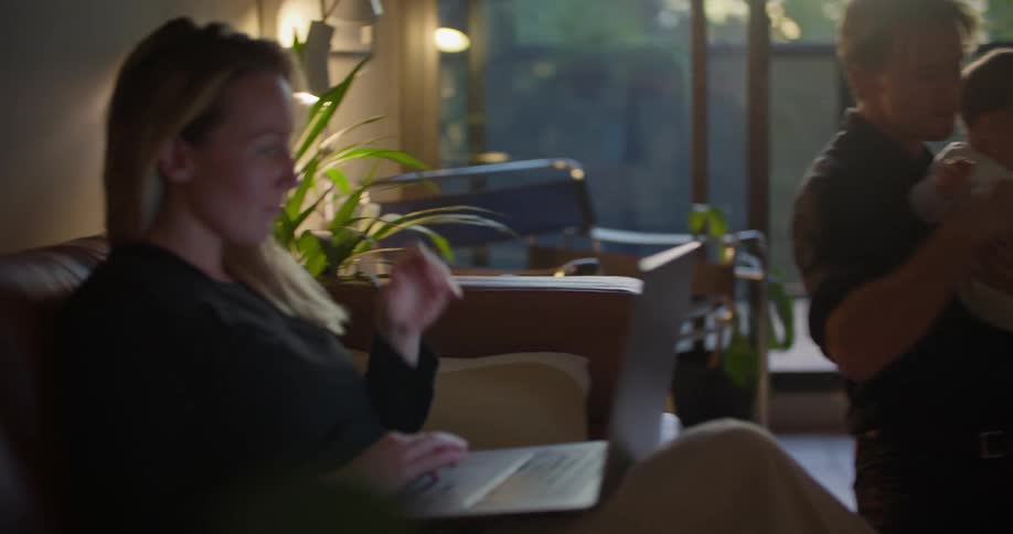 Father holds baby as mother sits on couch an uses laptop