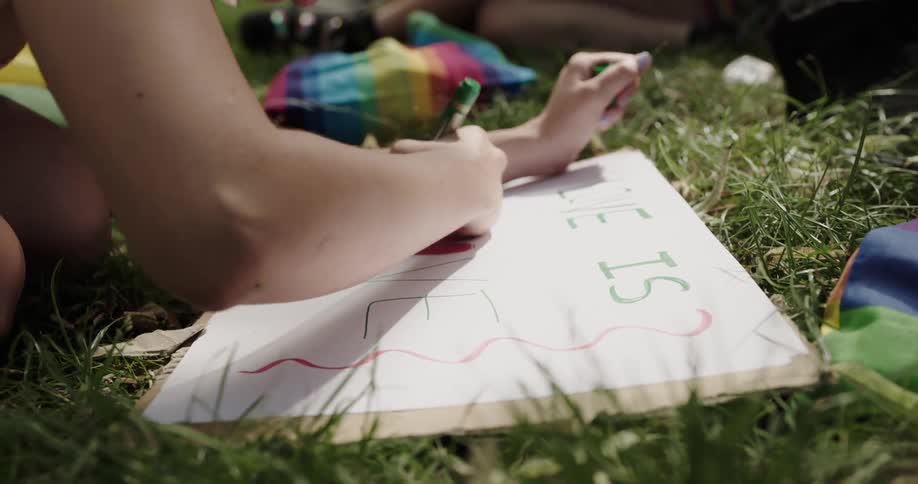 A woman makes a sign while sitting in the grass as her friends put on makeup behind her