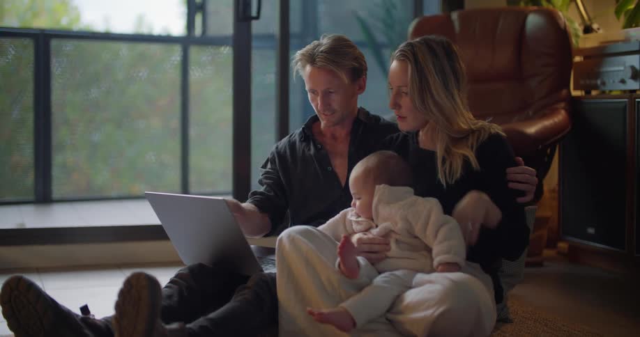 A man is sitting on the floor with a laptop computer and his wife is sitting next to him with their baby in her lap