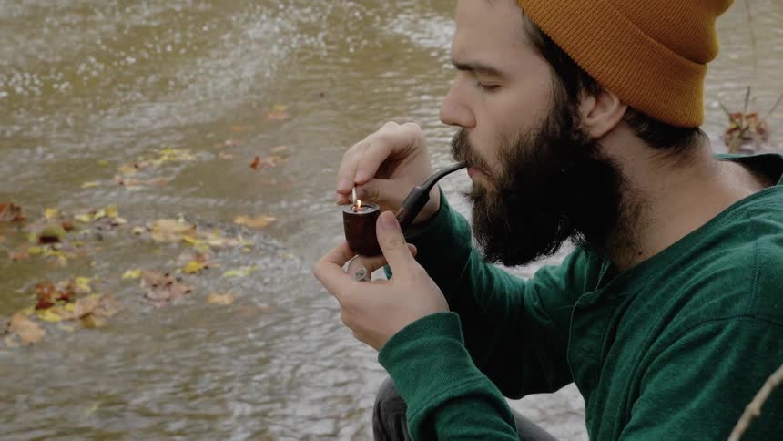 A man by a stream lights a pipe and smokes