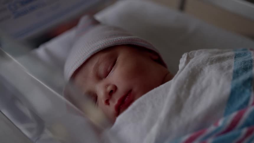Baby with pink hat and white blanket sleeps in bed