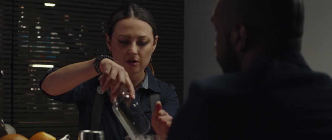 Female bartender preparing a drink by pouring ice into a glass while a man awaits sitting at the counter