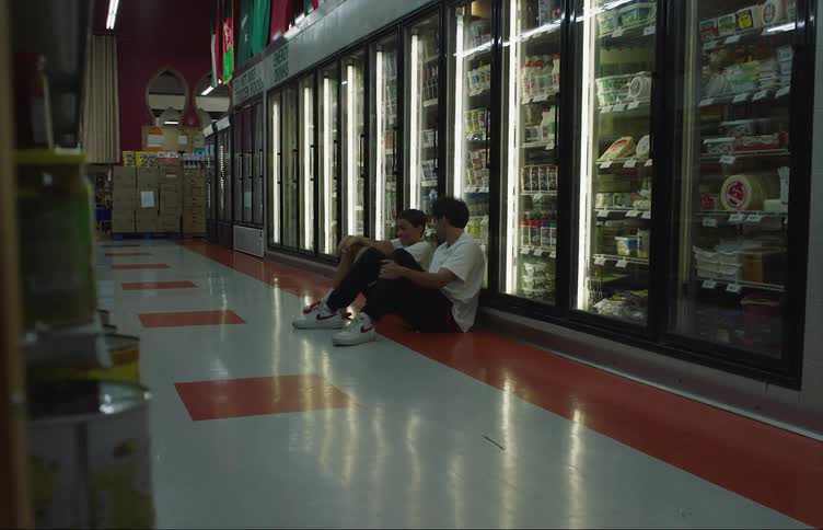 Two young men sit on the floor of a grocery store in front of frozen foods