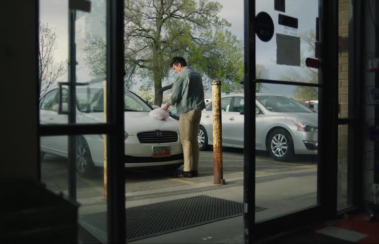 A man talking on a phone walks over to his parked car and opens the plastic shopping bag on the hood