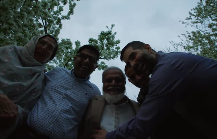A family hugs and leans over to look at the camera outdoors under some trees