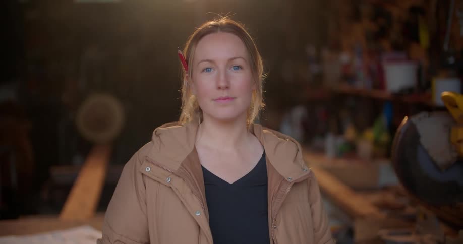 A woman is wearing a coat inside a wood shop and she puts a pencil behind her ear and smiles