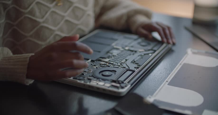 A woman is sitting at a desk and spinning a part on the inside of an opened laptop
