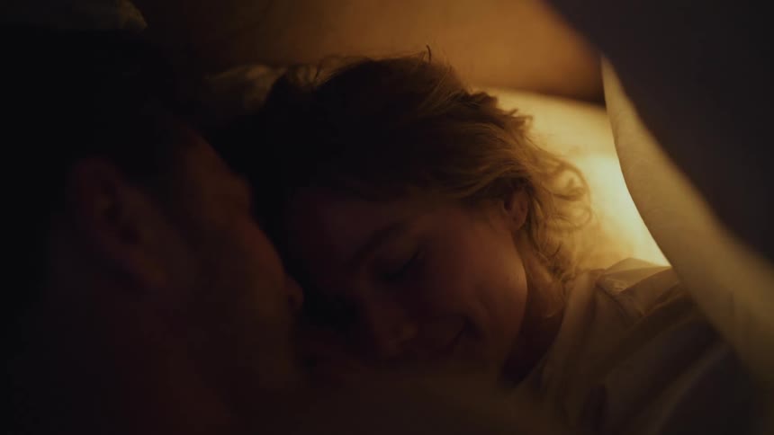 Woman with brown hair and man lie in bed smiling and cuddling in bedroom