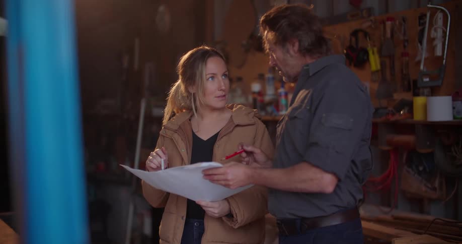 A woman references a large sheet of paper while talking to man standing beside her in a workshop during the day