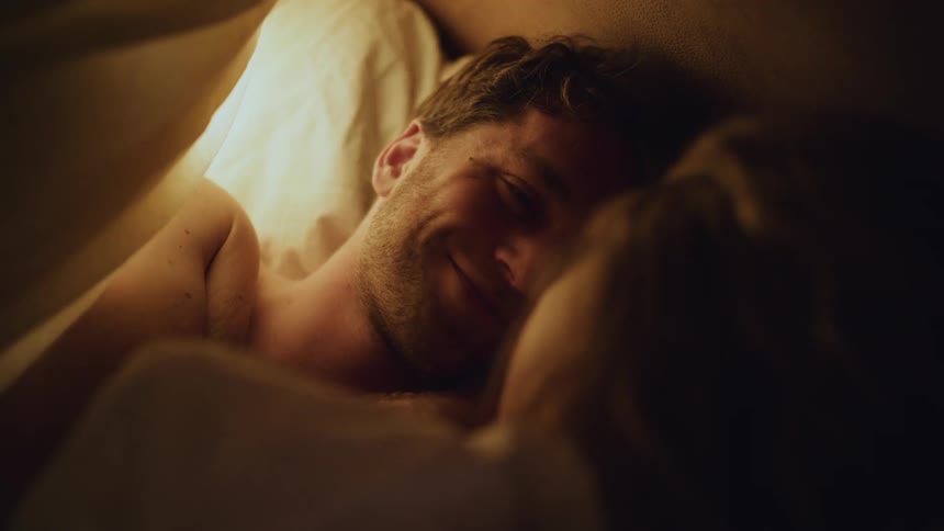 Couple lie in bed talking face to face in shadowy room under the sheets