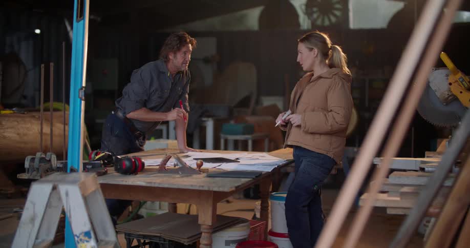 Woman and man discuss plans while standing in workshop with large tree trunk and power tools