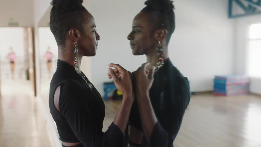 A woman walks across a room to examine herself in a large mirror covering a wall before turning to look into the camera