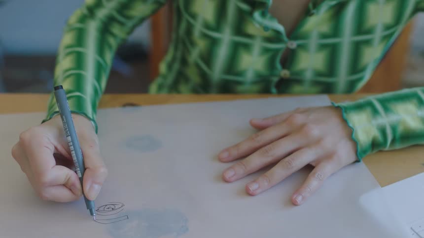 A woman draws on a sheet of paper while sitting at a table indoors during the day
