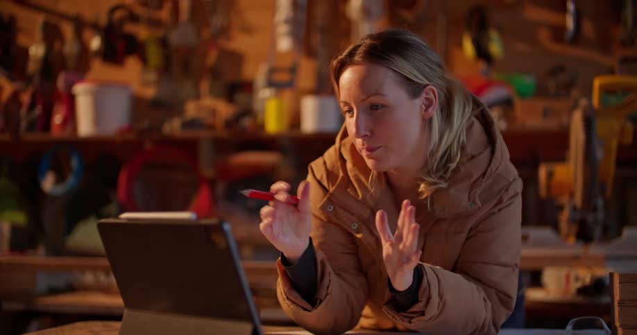 A woman is standing in a wood shop having a video conversation with someone on a laptop computer