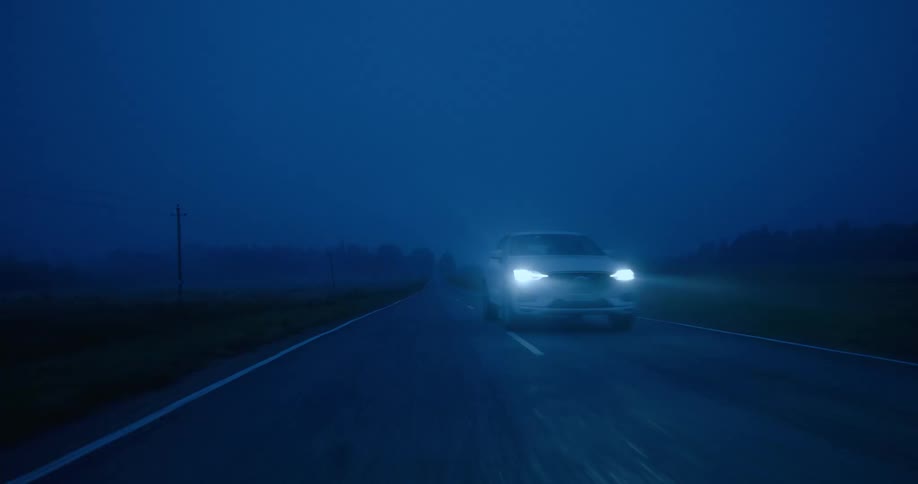 A white automobile with headlights on is traveling fast on a road and moves to the next lane