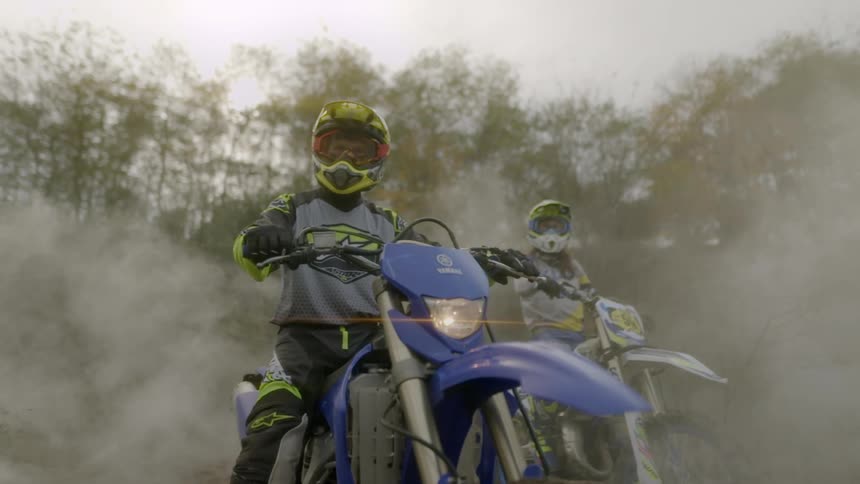 Two people are sitting on dirt bikes in a wooded landscape