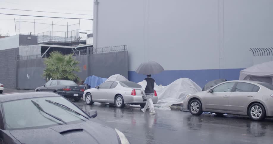 A man using an umbrella walks down a street lined with parked cars
