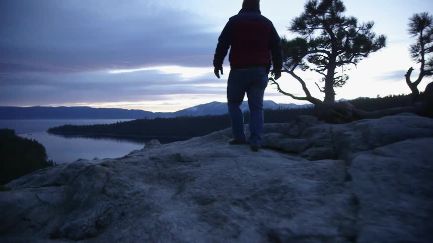 A man walks on a rocky ridge at dawn toward a view of a lake