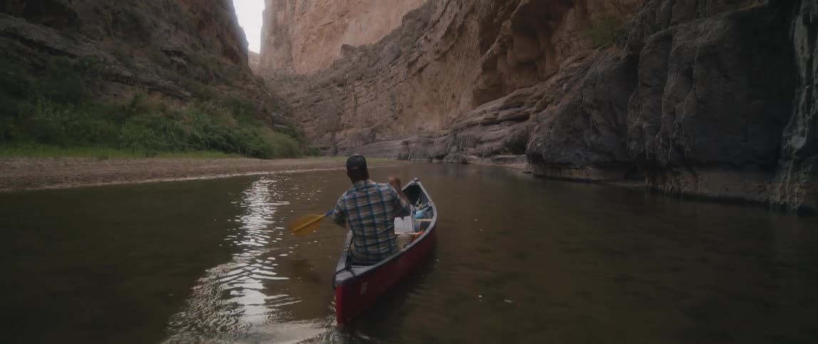 A man paddles a boat along a river on the floor of a canyon during the day