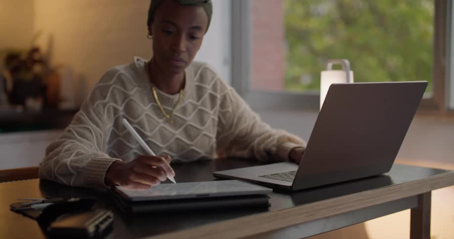 A woman is typing on a laptop and writing on a tablet as she sits at a desk