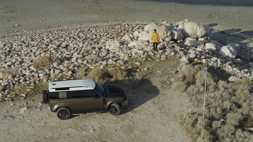 A man exists his vehicle and climbs atop a pile of boulders on a hill overlooking the coast on a foggy morning