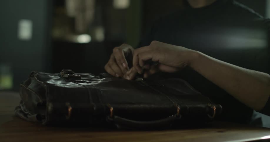 A man buckles the flaps on his leather briefcase and removes his glasses