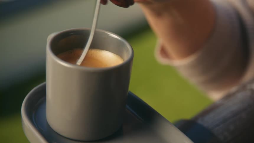 Womans hand using a coffee spoon to mix a small cup of coffee while shes sitting outside on a sunny day