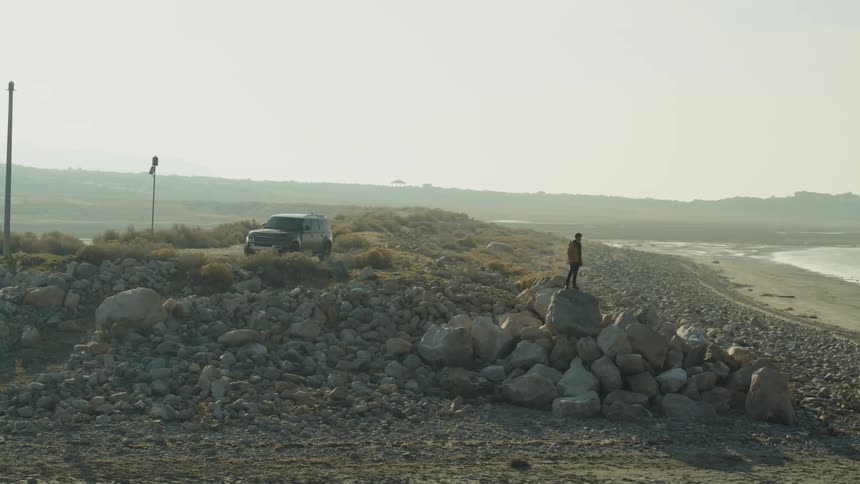 A man standing atop a pile of large stones on the coast takes a photograph of the ocean before climbing down and heading towards his vehicle