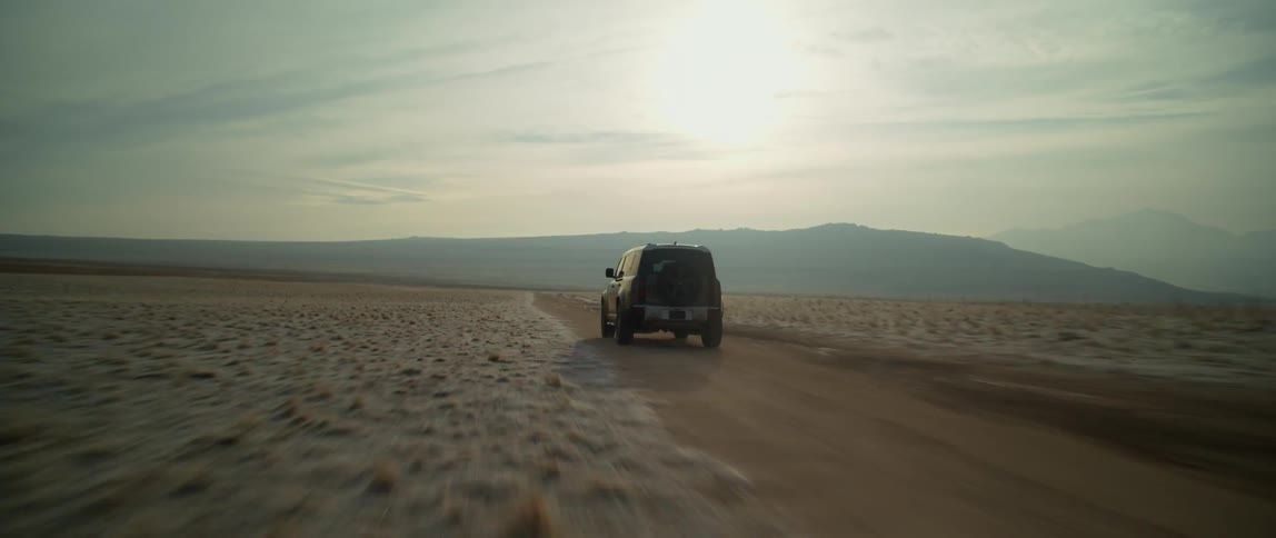 A black truck is traveling fast on a road across the desert and there are mountains in the distance